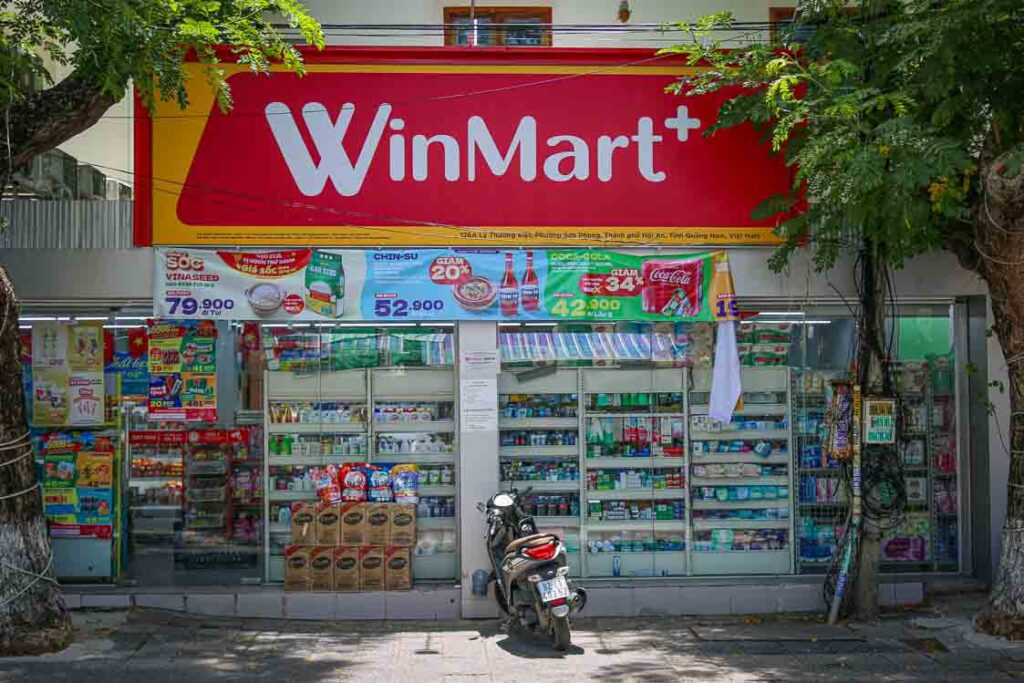 Exterior of a WinMart+ convenience supermarket in Hoi An with a large red sign above the entrance, glass-fronted displays, promotional banners and a motorbike parked outside.