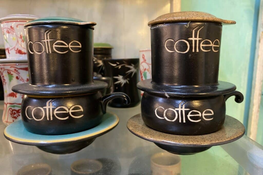 Two ceramic Vietnamese souvenir coffee drip makers with coffee written on them behind many other phin coffee filter souvenirs