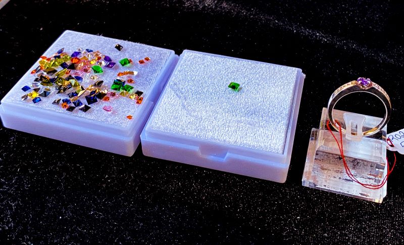 Coloured stones on small white display pad next to a middle pad with selected green stone on it. At the end is the ring to have stone replaced. This shows the process of custom-making your own ring at Ruby Jewellery Hoi An
