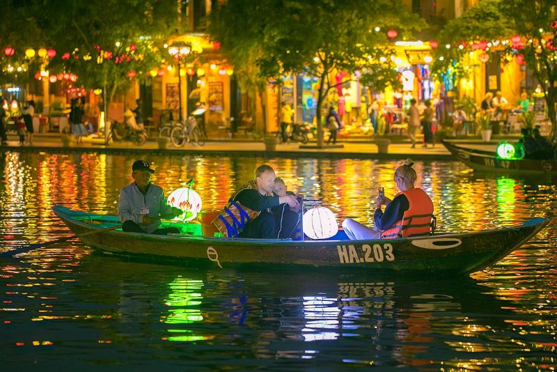 Tourists take a ride in a lantern-lit boat on the river with Hoi An Old Town equally lit with lanterns, in the background.