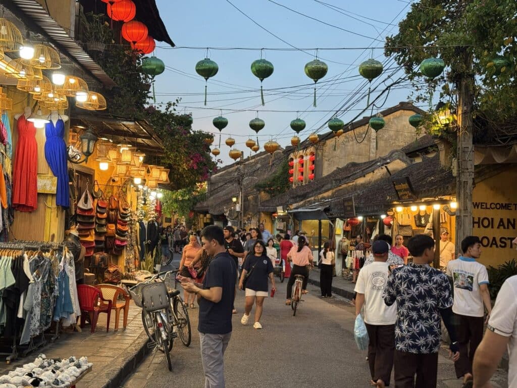 Tourists shopping along a busy street in Hoi An Ancient Town, surrounded by boutiques, leather shops, clothing stores and colourful lanterns.